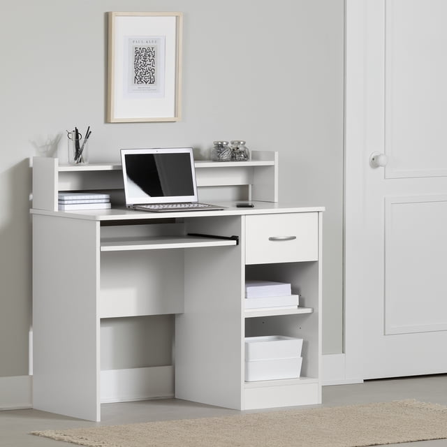 South Shore Smart Basics Desk in white for home office use, featuring a slide-out keyboard tray, one drawer with a silver handle, open shelving with books and storage bins, and a raised hutch shelf holding jars and stationery, set against a light grey wall with framed abstract art and a closed white door nearby.