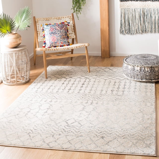 SAFAVIEH Tulum Glen Moroccan Geometric Area Rug in soft cream and grey hues with a subtle geometric pattern, laid on light wood flooring. It pairs with a woven wooden chair featuring a colorful patterned cushion, a black and white patterned pouf, a wicker side table with a wooden vase holding palm leaves, and a fringed wall hanging, creating a warm boho-chic atmosphere. Made with soft synthetic yarns, this durable area rug adds comfort and style to living spaces and is often used to enhance cozy reading nooks.
