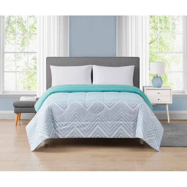 Mainstays Chevron Reversible Comforter Twin on a bed with a gray upholstered headboard in a bright bedroom. The comforter features a light blue and white chevron pattern microfiber polyester front and a solid turquoise reversible back. It is soft, cozy, and machine washable for easy care. The room includes white pillows, light wood flooring, a gray bench with books, a white nightstand with a blue geometric lamp, and large windows with white curtains.