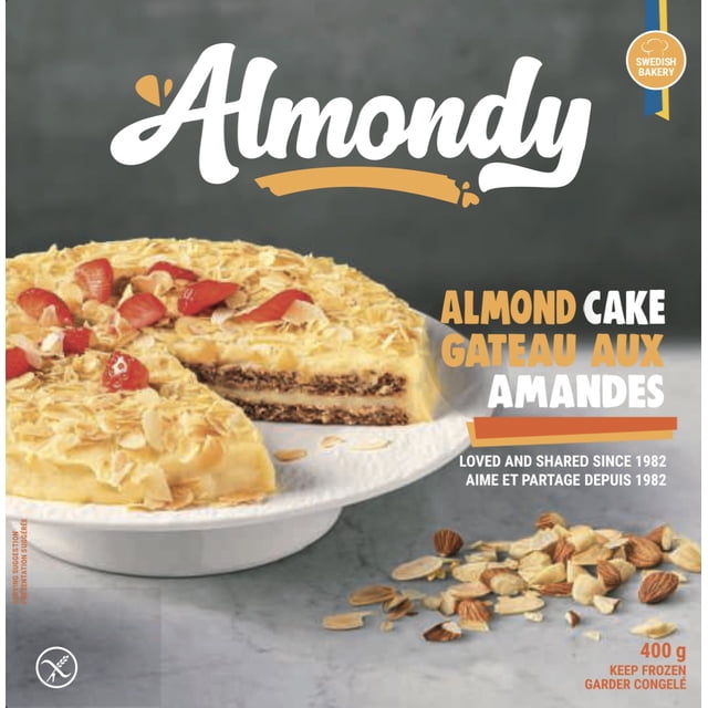 Gâteau aux Amandes sur une Assiette Piédestal Blanche avec une Tranche Enlevée, Couches Révélatrices de base aux Amandes et Crème Dorée Surmontées de Flocons d'Amande Rôtis et de Morceaux de Fraise, Entouré d'Amandes Dispersées sur une surface Grise, Convient comme Gâteau dessert Quotidien de la Marque de Boulangerie Suédoise Almondy.