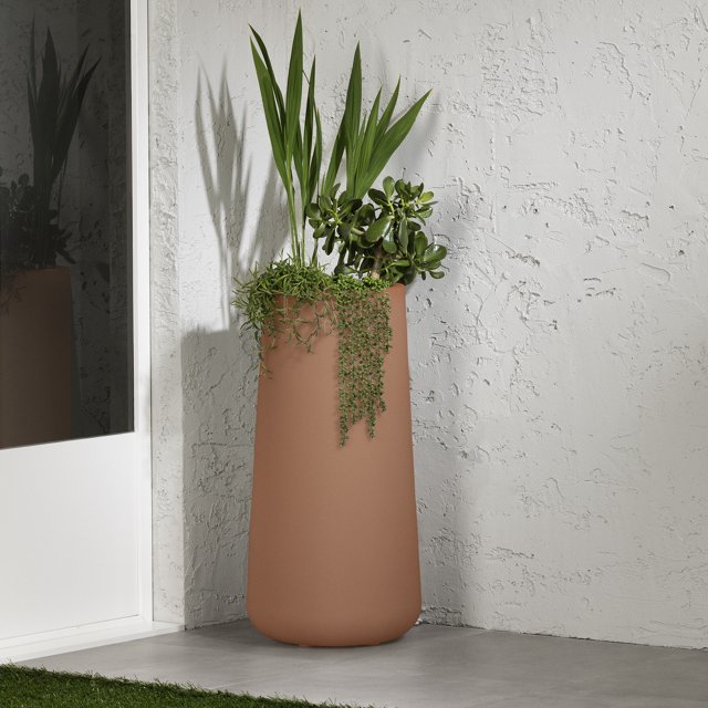 South Shore Dalya Tall Outdoor Pot Planter in terracotta, a large Scandinavian-style cylindrical planter made of durable polyethylene resin with a smooth matte finish, holding green plants including tall palm leaves and trailing succulents, placed on a concrete floor against a textured white wall near a glass door, ideal for enhancing outdoor garden or patio spaces.