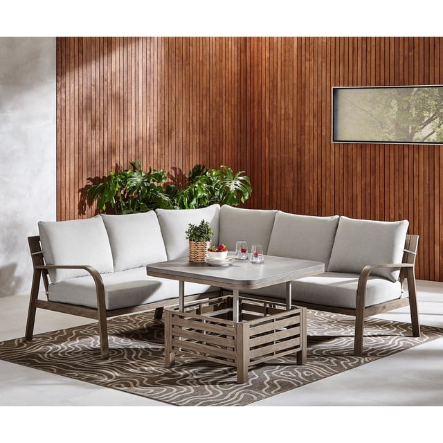 Better Homes & Gardens Prado 4-Piece Steel Outdoor Sectional Set with Cushions & Adjustable Table, Brown, shown indoors with a vertical wood-paneled wall. The sectional has brown rust-resistant powder-coated steel frames with a faux wood finish and light gray UV- and fade-resistant cushions. The adjustable steel table features a hand-painted concrete finish and holds a small potted plant, plates, and glasses. The set sits on a brown patterned rug with green plants behind it, suitable for outdoor or indoor patio use.