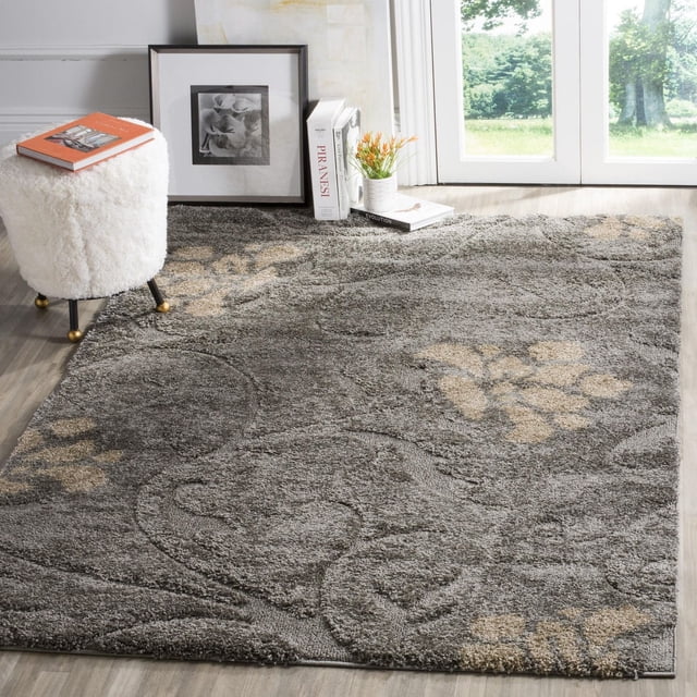 SAFAVIEH Florida Davin Floral Shag Area Rug with plush charcoal grey synthetic yarn and beige damask floral patterns, placed on light wood flooring beside a white faux fur stool, framed floral photo, books, and a small potted plant near a glass door, suitable for enhancing texture and comfort in living areas.
