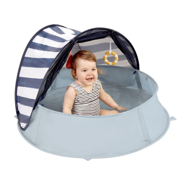Babymoov Aquani Tent & Pool with a light blue round waterproof pool base and a navy and white striped pop-up canopy providing UPF 50+ sun protection, including a soft mattress and hanging yellow and white ring toy, shown with a toddler sitting inside the water-filled pool.