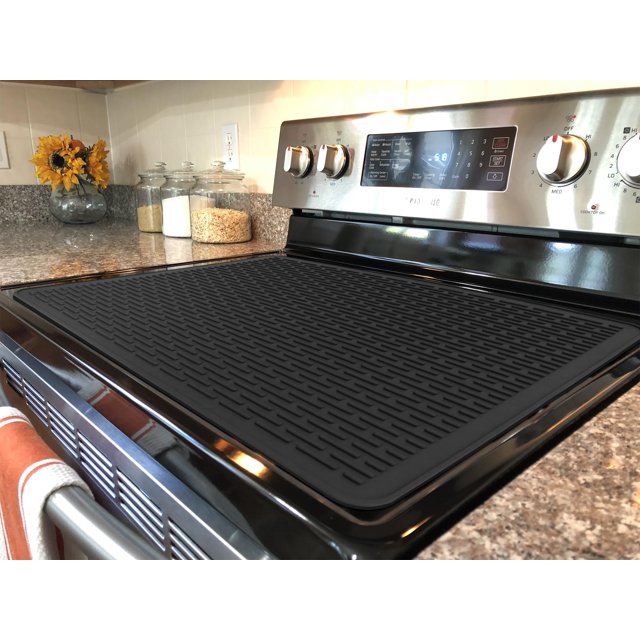 BreaDeep Silicone Electric Stove Cover Mat in black, measuring 28 x 20 inches, placed on a glass-top electric stove. The mat features wide raised line ridges for air circulation, heat resistance up to 450°F, and serves as a cooktop protector and expandable workspace. It protects the stovetop from scratches and spills while doubling as a drying mat or trivet. The kitchen setting includes a granite countertop, jars of grains, and sunflowers in a vase.