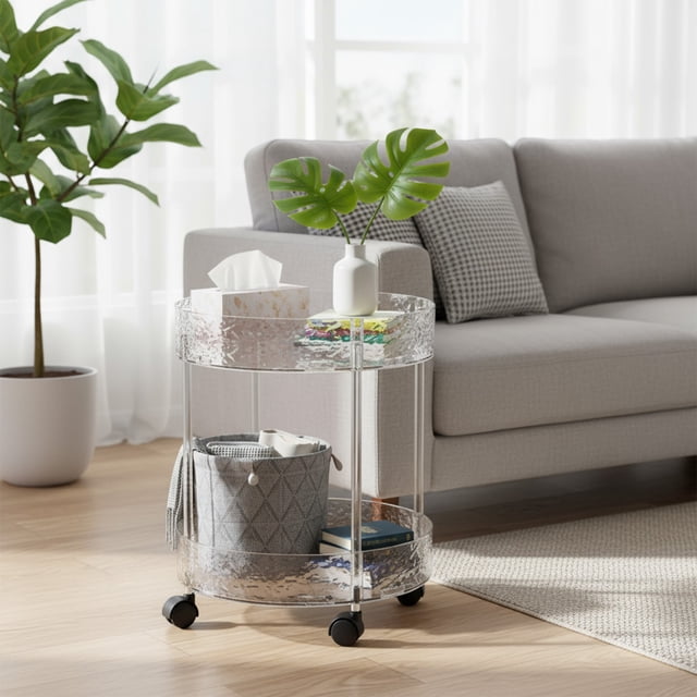 Rvkxad Two Tier Rolling Storage Cart, a multi-layer transparent movable trolley made of clear plastic, shown in a living room setting. The cart features two circular tiers with a textured transparent design, supported by a stable frame and four black caster wheels for easy mobility. The top tier holds a tissue box, a small vase with green leaves, and other small items, while the bottom tier contains a grey storage basket and books. Positioned on a wooden floor near a grey sofa and a potted plant, the cart demonstrates its versatility as a portable shelf organizer for various household items.