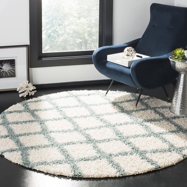 Safavieh Dallas Jerrie Geometric Shag Area Rug, round with a plush diamond pattern in cream and light grey, placed on a dark floor beside a navy blue armchair and a silver side table with a succulent, suitable for high-traffic home areas.