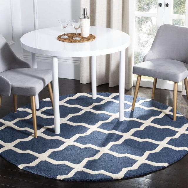 Safavieh Cambridge Liam Geometric Area Rug, round with a blue base and white geometric lattice pattern, hand-tufted wool texture, placed under a white round dining table with two grey velvet chairs with gold legs on dark hardwood floors near a window with beige curtains, ideal for adding modern American style and warmth to home dining areas.
