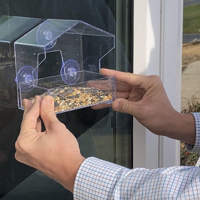 WINDOW BIRD FEEDER with clear plastic design and three suction cups being attached to a window, featuring a removable seed tray filled with mixed birdseed, ideal for close-up bird watching in outdoor garden areas.