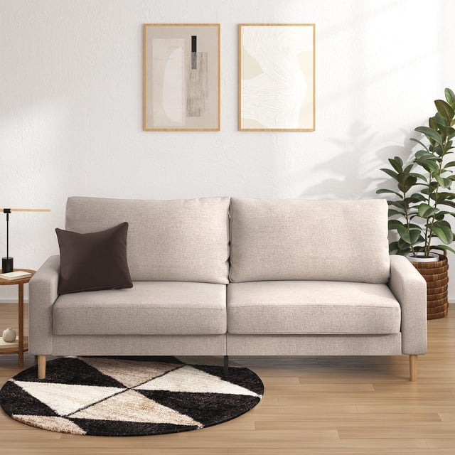 Zinus Colton 76 inch 3 Seat Upholstered Sofa Couch in beige fabric with squared arms and tapered wood legs, front-facing on a light wood floor with a geometric round rug, next to a wooden side table with lamp, abstract wall art, and a potted plant, suitable for modern living rooms.