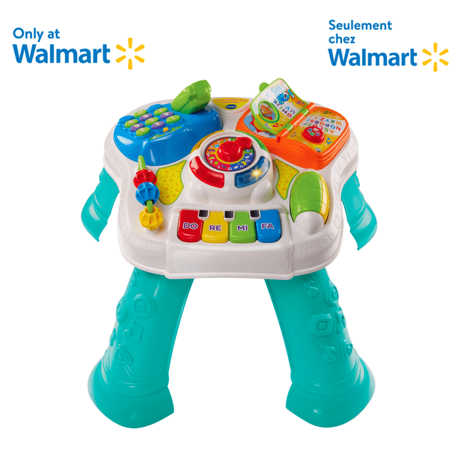VTech Sit-to-Stand Learn & Discover Table, a multicolour educational electronics toy for babies with a white activity panel and teal legs, featuring six interactive stations including a blue phone with number pad, an orange alphabet book, a central clock with red dial, colorful piano keys labeled Do, Re, Mi, Fa, and sliding beads, designed for floor play or as a standing activity table.