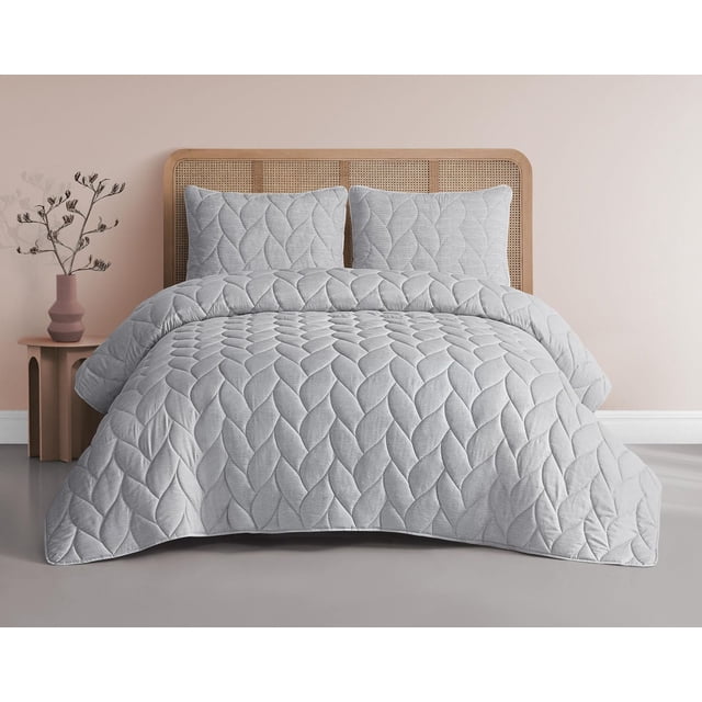 Emily 3 Piece Reversible Comforter Set in grey displayed on a bed with a woven wooden headboard, featuring a braided quilt pattern on the microfiber comforter and two matching shams, set in a minimalist bedroom with a pink wall and a side table holding a vase with dried branches, machine washable and reversible for versatile use.