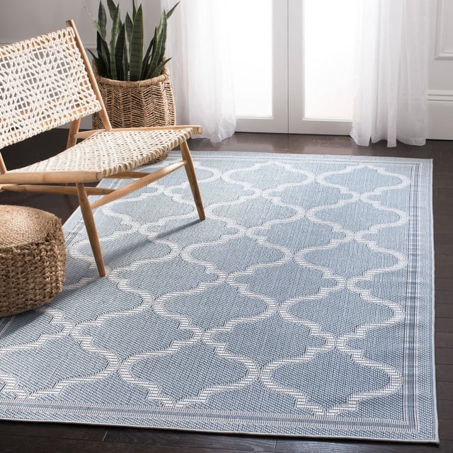 SAFAVIEH Bermuda Corie Geometric Quatrefoil Outdoor Area Rug in light blue with white quatrefoil pattern on dark hardwood floor, next to a woven rattan chair and basket planter with snake plant, illuminated by natural light through sheer white curtains, ideal for outdoor patios or indoor sunrooms.