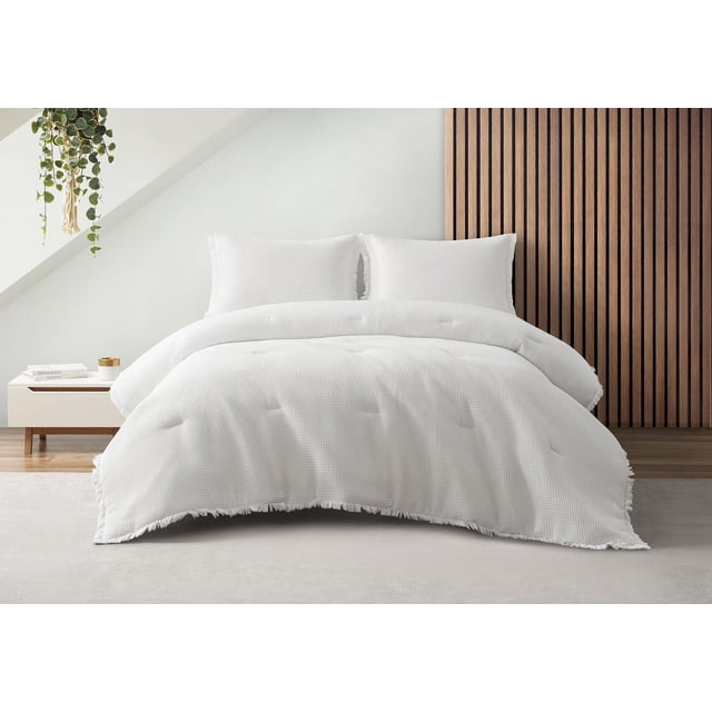 Charlotte 3 Piece Waffle Cotton Comforter Set in white on a bed with two matching shams, featuring a textured waffle cotton fabric, fringe trim on the comforter edges, and tassels on the sham sides, displayed in a modern bedroom with wood slat wall and hanging plant, ideal for adding warmth and softness to bedding.