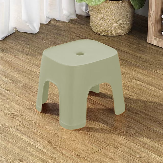 Fridja Non Slip Step Stool Shower Foot Stool, 8 inches high, made of thickened plastic in light gray. The stool measures 8.07x7.67x6.29 inches and features a curved ergonomic design for comfort, a slip-resistant textured base, and reinforced legs for stability. Suitable for home, office, garage, or bathroom use. Lightweight and portable, ideal for various tasks such as stepping, sitting, or as a children's stool. Shown in a home setting on a wooden floor with a plant and storage unit in the background.