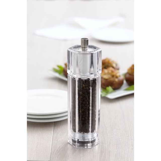 Trudeau 6.5" Octo Combo Pepper Mill/Salt Shaker with clear acrylic six-sided body filled with black peppercorns, featuring a ridged metal top knob for adjustable grind from coarse to fine, standing on a light wood table with blurred white plates and stuffed mushrooms in the background.