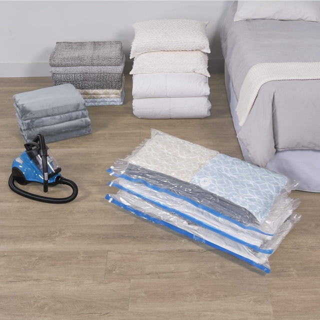 Mainstays Ensemble de 3 Sacs sous Vide Jumbo Compressés sous le Lit sur un Plancher de Bois à Côté d'Une Pompe à Vide Bleue, avec Couettes Pliées, Oreillers et Couvertures Empilés à Proximité et un Lit avec une Literie Grise en Arrière-Plan, Montrant un Rangement Gain de Place pour les Draps Volumineux et les Manteaux d'Hiver.
