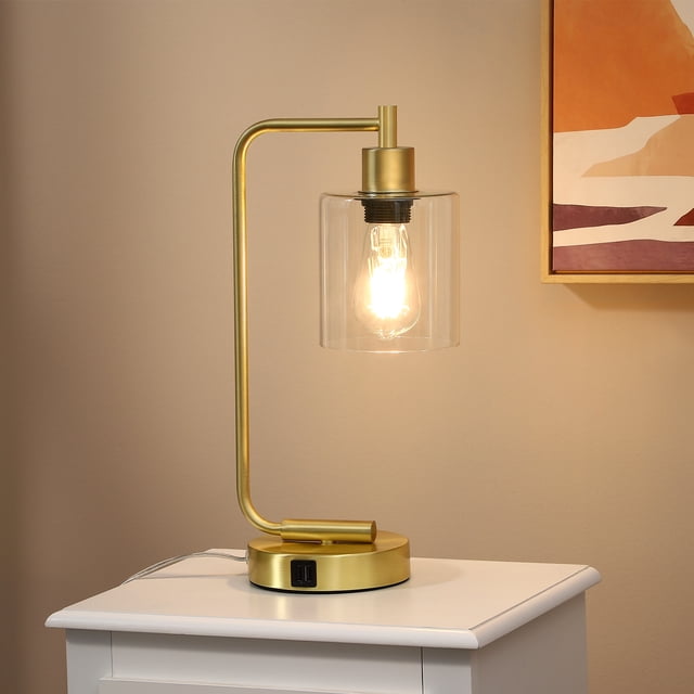 EDISHINE Gold Lamps Table, a small bedside lamp with a sleek industrial style featuring a gold curved arm and pedestal base. The lamp includes a clear cylindrical glass lampshade showcasing the included dimmable 3000K LED bulb. It offers touch control functionality, three brightness settings, and dual USB charging ports on the base for convenience. Suitable for bedroom or desk use.