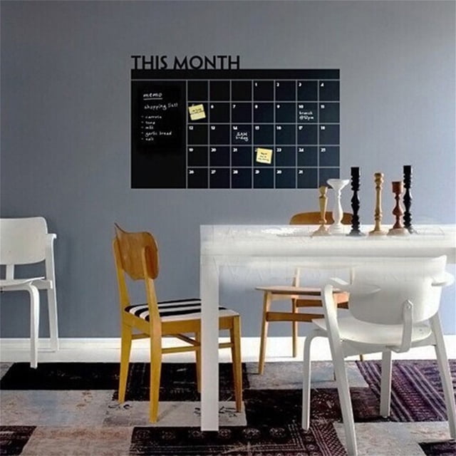DPTALR 60x92 Month Plan Calendar Chalkboard Blackboard Vinyl Wall Sticker displayed on a smooth grey wall in a modern dining area. The blackboard features a monthly calendar layout with grid lines and sections for notes, along with sticky notes for added organization. The setting includes a white dining table surrounded by wooden and white chairs, with decorative candlesticks on the table. The wall sticker is made of PVC material, removable, and suitable for smooth surfaces like walls, tiles, or glass.