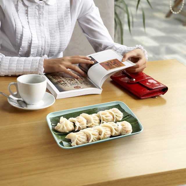 Fridja Summer Savings Rectangular Plates Hot Pot Barbecue Plastic Sausage Plates Fruit Plates. A rectangular light green plastic plate with a square-inspired design, holding dumplings on a banana leaf. The plate is on a wooden table with a white coffee cup, a red wallet, and an open book, ideal for versatile dining and entertaining needs.
