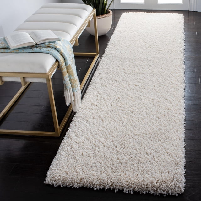 Safavieh California Solid Plush Shag Area Rug in off-white with a dense 4.5kg/m² polypropylene pile, placed on dark hardwood flooring beside a white cushioned bench with gold metal frame, accented by a blue and gold throw and open book, near a potted plant and glass doors, suitable for adding softness and warmth to home interiors.
