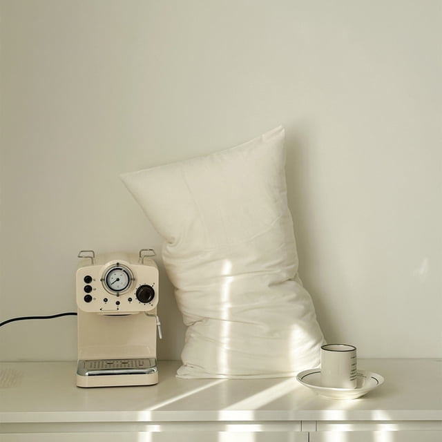 RKXZT Standard Cotton Pillowcases with envelope closure in a cream color, measuring 20x30 inches. These breathable and soft bed pillow cases are made of natural cotton, ideal for bedrooms, hotels, and dormitories. Displayed in a minimalist setting with a coffee machine and cup nearby, they complement soft bedding decor.