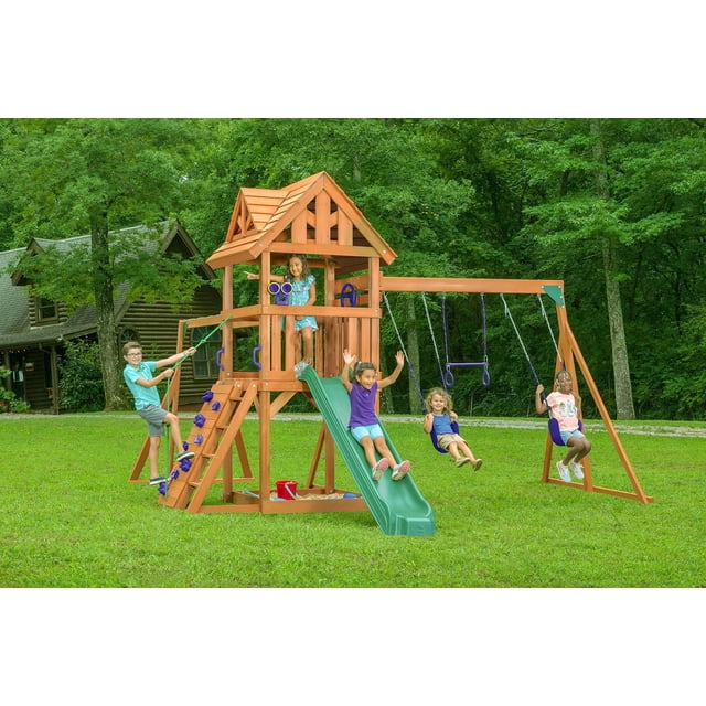 Ensemble de Jeu en Bois Style Chalet avec Toit en Bois Comprenant une Tour en Cèdre avec une Terrasse de 4 Pieds, Glissade Verte, Mur d'Escalade, Échelle en Bois, Balançoires Côte à Côte, Barre de Trapèze et Bac à Sable en Dessous, Installé sur une Cour Arrière Gazonée Près d'Une Cabane en Rondins et Entouré d'Arbres, Conçu pour les Enfants de 3 à 10 Ans.