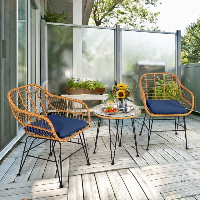Costway 3PCS Patio Rattan Bistro Furniture Set Cushioned Chair Table Navy arranged on a wooden patio. This outdoor bistro set includes two rattan chairs with navy seat cushions and curved armrests, along with a round coffee table featuring a tempered glass top. Supported by black steel frames with anti-slip foot pads, the set is ideal for outdoor spaces like patios or balconies. Sunflowers and refreshments on the table add a welcoming touch.
