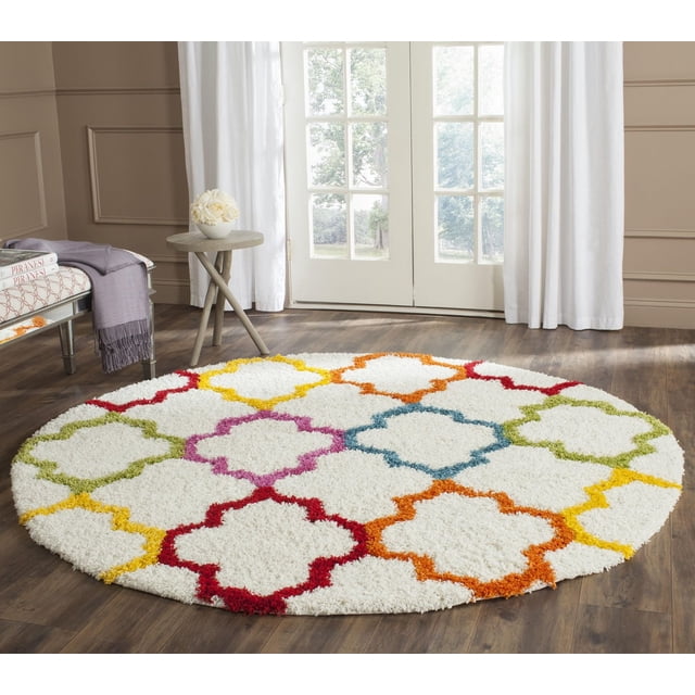 Safavieh Shag Kids Yadira Geometric Quatrefoil Area Rug, round with a white 100% wool pile featuring colorful geometric quatrefoil patterns in red, orange, yellow, green, blue, and pink, placed on a hardwood floor in a bright room with white curtains and glass doors, ideal for adding vibrant style to children's bedrooms or playrooms.