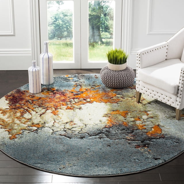 Safavieh Glacier Nazca Abstract Area Rug, round with a marbled abstract pattern in blue, orange, cream, and gray tones, placed on dark hardwood flooring beside a white studded armchair, two white vases with blue stripes, and a gray knitted pouf with a green plant, near glass doors showing a green outdoor landscape. This durable accent rug is also suitable for adding artistic flair to reading nooks or lounge areas.