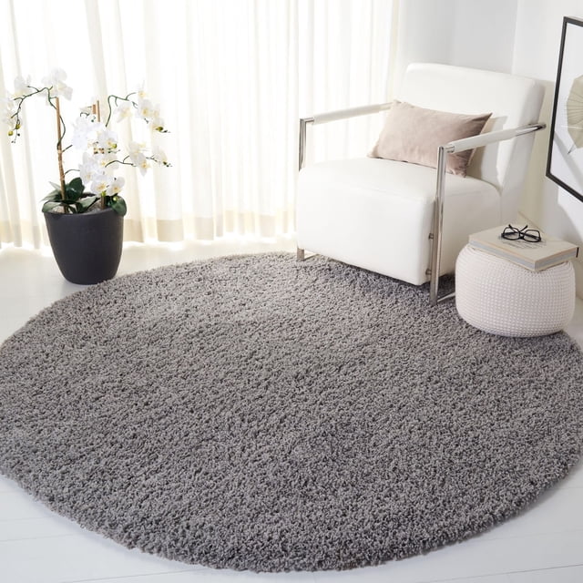 Safavieh Venus Elijah Solid Plush Shag Area Rug, round with a 1.77-inch medium pile in grey, placed on a light floor beside a white armchair with metal arms and a beige pillow, a white knitted pouf with books and glasses, and a black planter with white orchids in a bright room with sheer curtains, ideal for adding plush texture and warmth to contemporary living spaces.