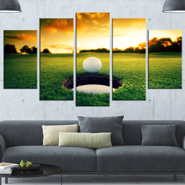 Design Art Golf Ball near Hole Landscape Artwork Canvas Print showing a five-panel gallery wrapped canvas with a close-up of a white golf ball on the edge of a golf hole on green grass under a warm sunset sky, ready to hang above a modern grey sofa in a minimalist living space.