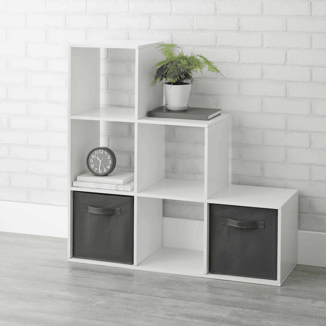 Mainstays 3-2-1 Cube Storage Organizer in white with six open-back cube shelves arranged in a stair step design, featuring two black foldable storage bins in the bottom compartments, a small round clock and books in the middle left cube, and a potted fern with a closed book on the top right cube, set against a white brick wall on a light wood floor. Suitable for organizing books, decor, and small household items.