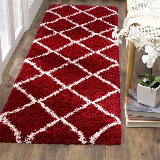 Safavieh Hudson Amias Geometric Shag Area Rug in deep red with white diamond lattice pattern, featuring a thick plush shag pile designed for durability in high-traffic areas, placed on wood flooring beside a white woven chair and a stack of books with a white orchid plant, often used to add texture to modern or urban décor.