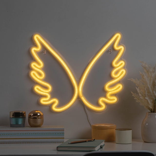 Merkury Innovations 15.75" Wings LED Neon Wall Sign with warm yellow neon effect mounted on a grey wall above a desk featuring decorative jars, a notebook, pen, and a vase with dried pampas grass, USB powered with included 6-foot cable, suitable for adding unique novelty lighting to home interiors.