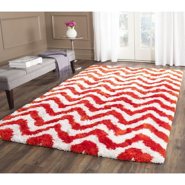 Safavieh Barcelona Carmella Zigzag Stripes Shag Area Rug with plush shag texture in red and white zigzag stripes, displayed on dark hardwood flooring in a bright room with beige walls, a gray upholstered bench, and a round side table with a white vase of pink flowers, suitable for adding warmth and style to accent rug areas.