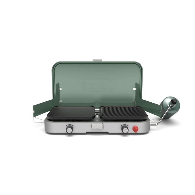 Coleman Cascade 3-in-1 Propane Camping Stove with open green lid, showing two burners with removable cast-iron grill and griddle accessories, silver base with two black control knobs and a red ignition knob, and a green propane regulator on the right side, suitable for versatile outdoor cooking.