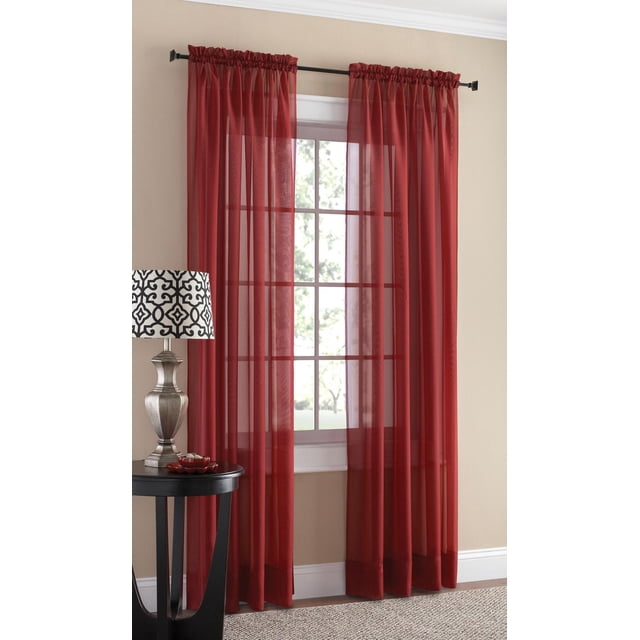 SUN TEXTILE VOILE ROD POCKET CURTAIN in red sheer voile fabric with built-in rod pockets hanging on a black curtain rod over a white-trimmed window, softly filtering light while enhancing privacy, displayed in a beige room with a black side table and silver lamp with patterned shade, suitable for layering on a double rod.