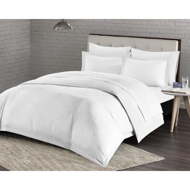 Millano Spa Ensemble de Housse de Couette en Microfibre Blanche 1200 Fils, Exposé sur un Lit avec une Tête de Lit Capitonnée beige. l'Ensemble Comprend une Housse de Couette et des Couvre-Oreillers, Doté d'Un Tissu Infroissable, Résistant à la Décoloration et Hypoallergénique. le Lit Repose sur un Tapis Texturé beige avec un Plancher en Bois et une table d'Appoint Ronde Tenant un Verre d'Eau et un Livre Ouvert, Ensemble contre un Mur de Briques Gris. Cet Ensemble de Literie Convient Aussi pour Créer un Environnement de Chambre d'Amis