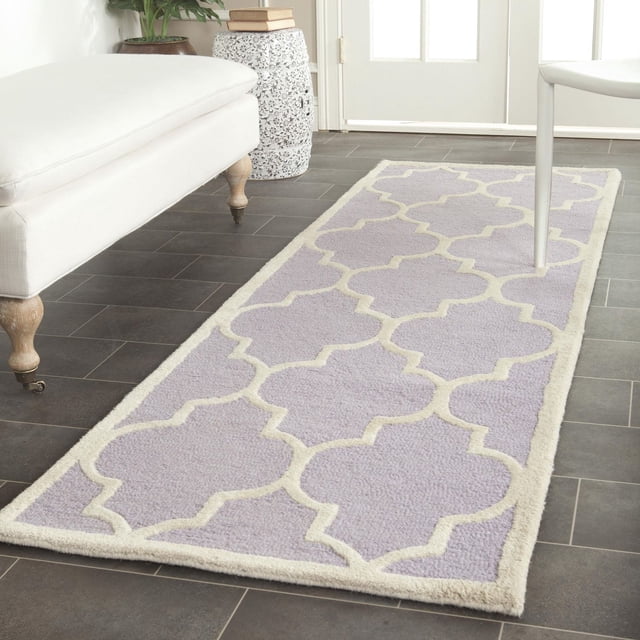 Safavieh Cambridge Kevin Geometric Area Rug with a lavender base and cream geometric trellis pattern, hand-tufted wool featuring plush cut and high/low loop pile, placed on dark tile flooring between white upholstered bench and chair near a door, ideal for adding modern American style to home or office.