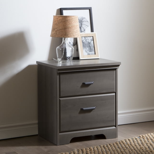 South Shore Versa Table de Chevet à 2 Tiroirs en Érable Gris avec Deux Tiroirs, avec Poignées en Métal et base à Découpe à Patte d'Éléphant, Surmontée d'Une Lampe en Verre Transparent avec Abat-Jour en Liège et Deux photos Encadrées en Noir et Blanc, Installée sur un Plancher de Bois Franc Près d'Un Mur Lumineux et d'Un Tapis Tissé, Convient au Rangement et à l'Organisation de la Table de Chevet.