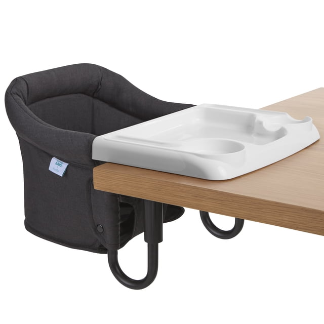 Evolur Clamp On Comfort Table Chair in black clamped to a light wood table edge, with a soft fabric seat and removable white plastic tray featuring cup holder and snack compartments, designed for toddlers aged 6-36 months for portable mealtime seating.