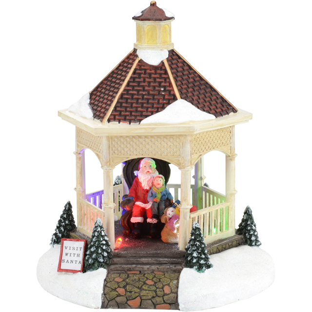 Light-up multi-color Christmas village gazebo featuring Santa Claus seated inside with children, surrounded by snow-covered pine trees and a 'Visit With Santa' sign.