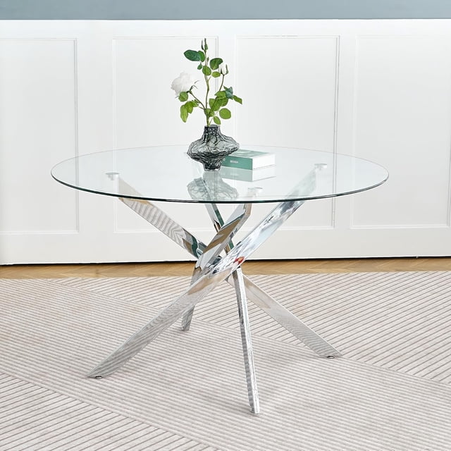 Heavenly Collection Star Dining Table with a 48-inch round clear tempered glass top featuring beveled edges and a polished chrome X-frame stainless steel base, displayed on a light striped rug with a black textured vase holding white roses and a design book on the tabletop, ideal for contemporary dining areas.