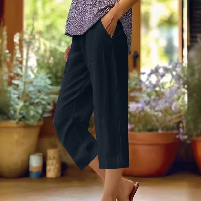 DPTALR Women's Summer Cotton And Linen Solid Color Pockets Elastic Waist Capris Shorts Pants in a breathable and lightweight polyester material. The black capris feature a solid color design, elastic waistband, and side pockets, offering a comfortable and casual fit. Suitable for lounging at home or outdoor activities, shown in a relaxed indoor setting with potted plants in the background.
