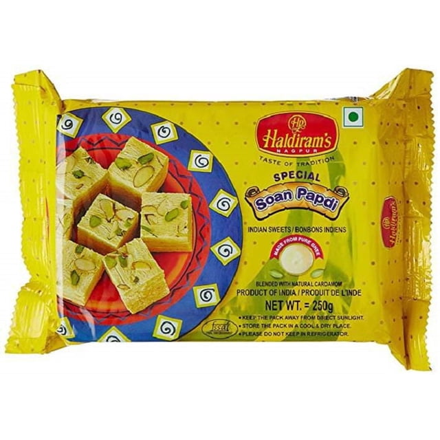 Haldiram Soan Papdi in yellow packaging displaying square pieces of flaky Indian sweet topped with pistachio slices on a decorative blue and red plate, labeled special Soan Papdi with natural cardamom, net weight 250g, an Indian mithai ideal for snacks and desserts.