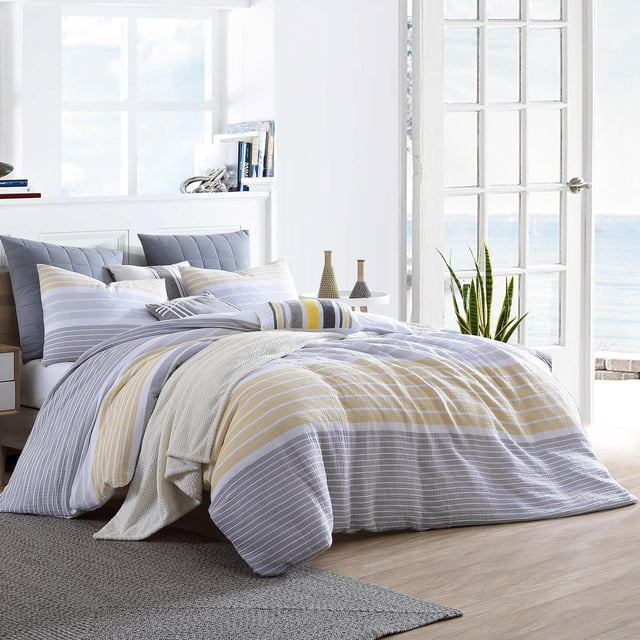 Ensemble de Housse de Couette en Coton – Collection Cordelia sur un Lit avec Tissu en Coton Rayé Jaune, Gris et Blanc et Taies d'Oreiller Assorties dans une Chambre Lumineuse avec Grandes Fenêtres Surplombant l'Océan, Convient pour Ajouter Confort et style à la Literie.