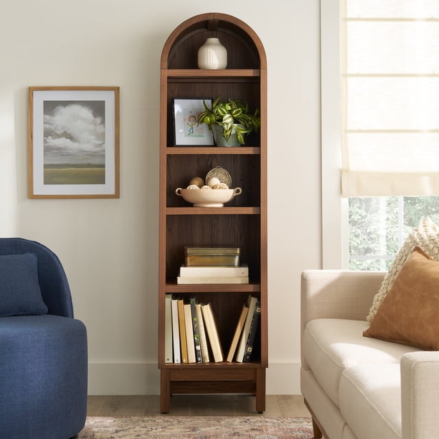 Better Homes & Gardens Juliet Bibliothèque Étroite en Arche, Fini Noyer, avec Cinq Tablettes, Dont Trois Ajustables, Orientée vers l'Avant avec Dessus Arqué, Tenant un vase Blanc, Oeuvre Encadrée, Plante en Pot, Bol Décoratif avec Boules Tressées et Livres. Positionnée entre un Fauteuil Bleu et un sofa beige dans un Salon avec Murs Légers et Stores de Fenêtre, Idéale pour le Mobilier de Bureau Ou la Déco de la Maison.