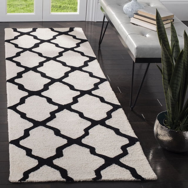 Safavieh Cambridge Liam Tapis Géométrique pour Aire en Vue Frontale sur Plancher de Bois Franc Sombre, Présentant de la Laine Tuftée à la Main avec un Motif de Treillis Géométrique Noir sur Blanc, Placé à Côté d'Un Banc Gris avec des Livres et un vase, Près d'Une Plante Serpent dans un pot Métallique et des Portes en Verre.