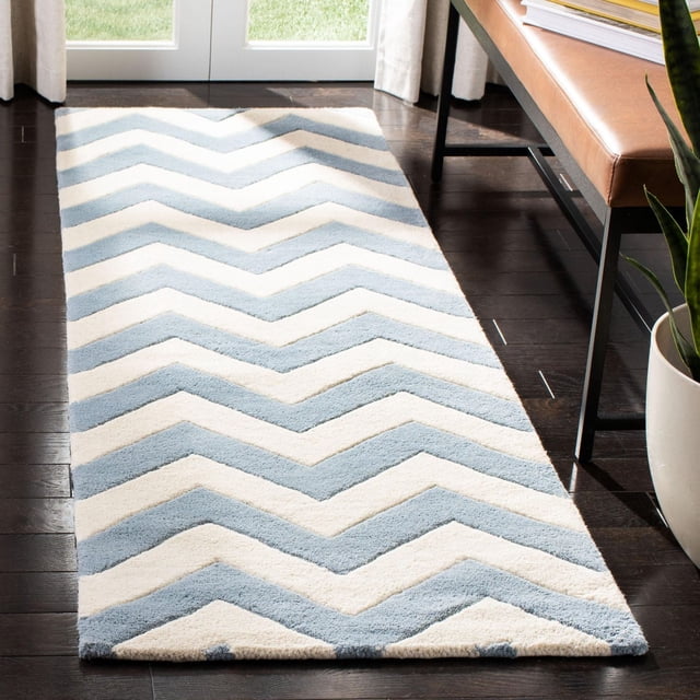 Safavieh Chatham Jake Zigzag Stripes Area Rug featuring hand-tufted wool with a plush medium pile, displaying a light blue and cream zigzag stripe pattern on dark hardwood flooring beside a leather bench and potted plant, suitable for contemporary accent rug use.